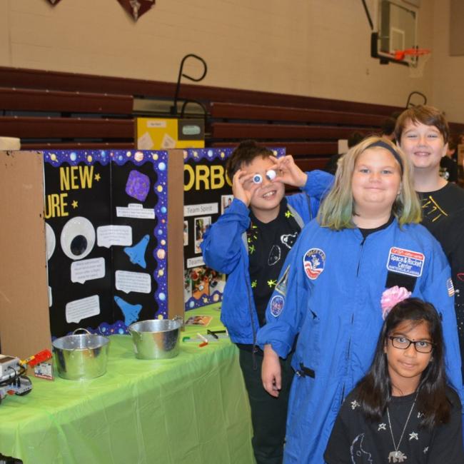 Lego League team poses for picture next to their project board and wearing space gear